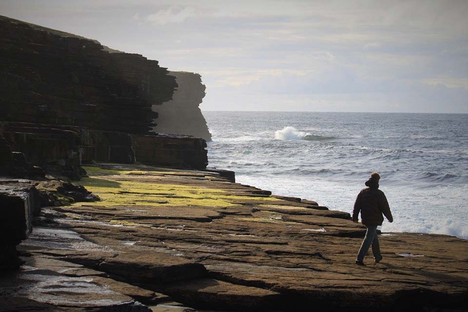 Student walking on Bristol coast as waves crash against cliff face