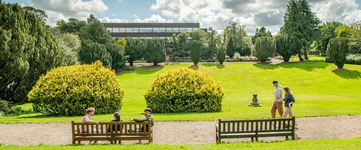 A group of students sitting on benches overlooking the lawn surrounded by trees and bushes.